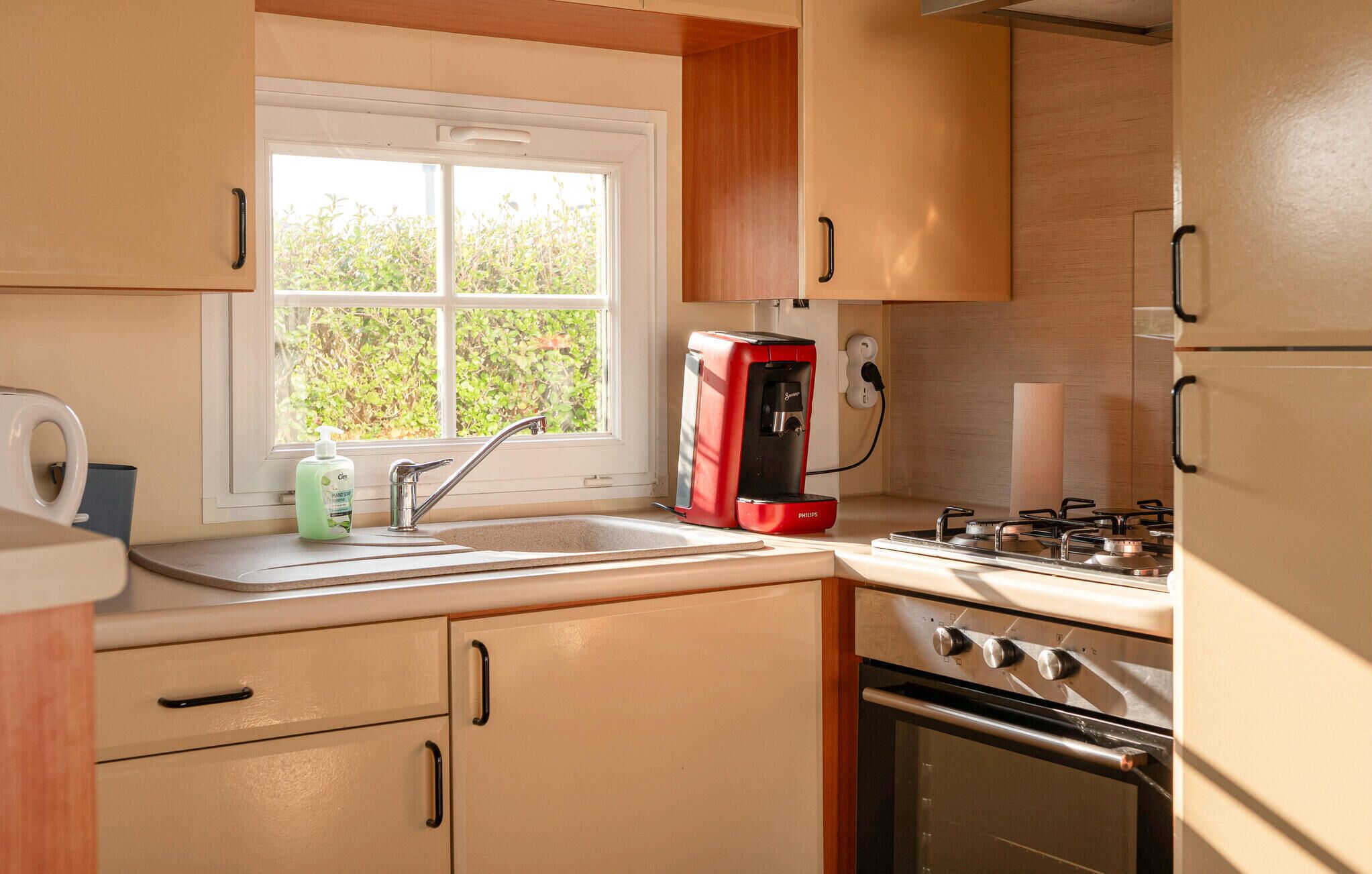 A compact kitchen with a sink, a red coffee maker, a stove, and wooden cabinets.
