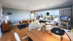 A modern living room with a dining area, large windows with a city view, and a television on a stand.