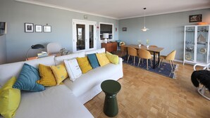 A living room with a white sofa, yellow and blue cushions, a dining table with chairs, a small cabinet with nautical decorations, and a wooden floor.