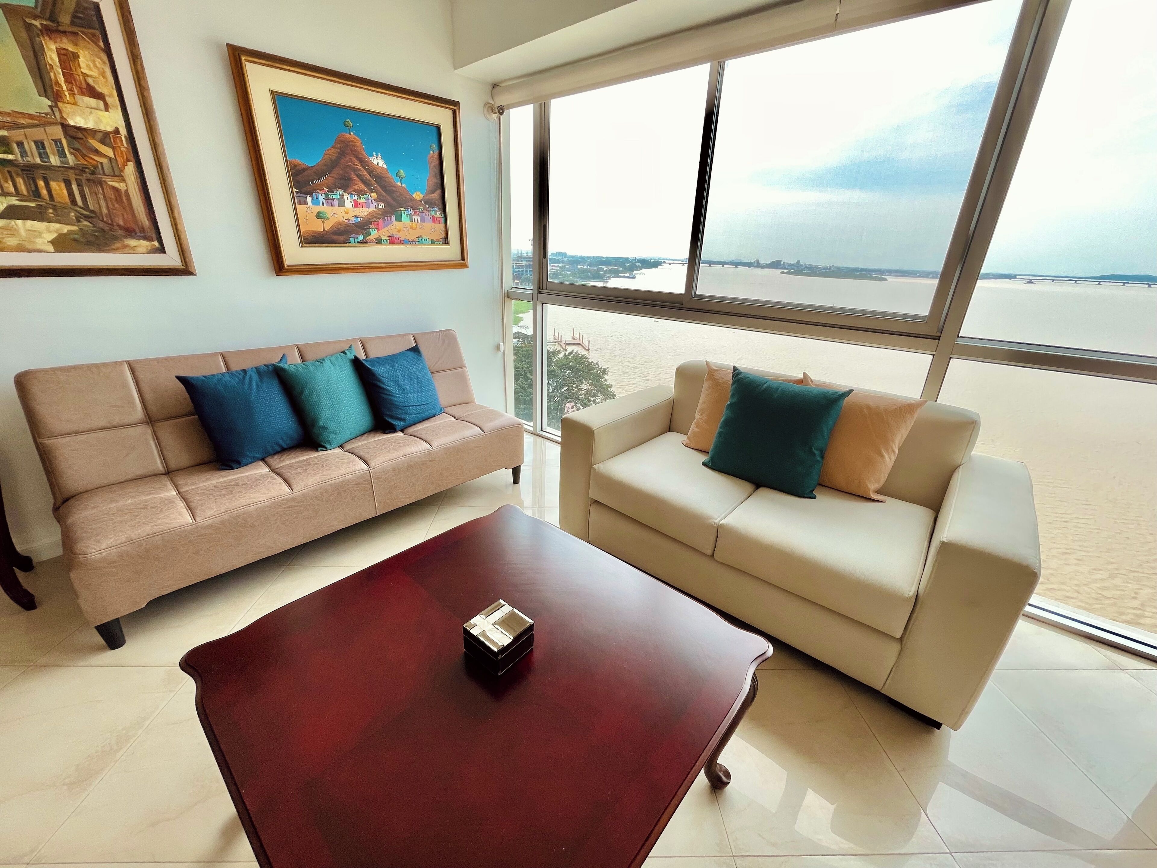 A living room with two sofas, a coffee table, and a large window offering a view of a river and distant cityscape.
