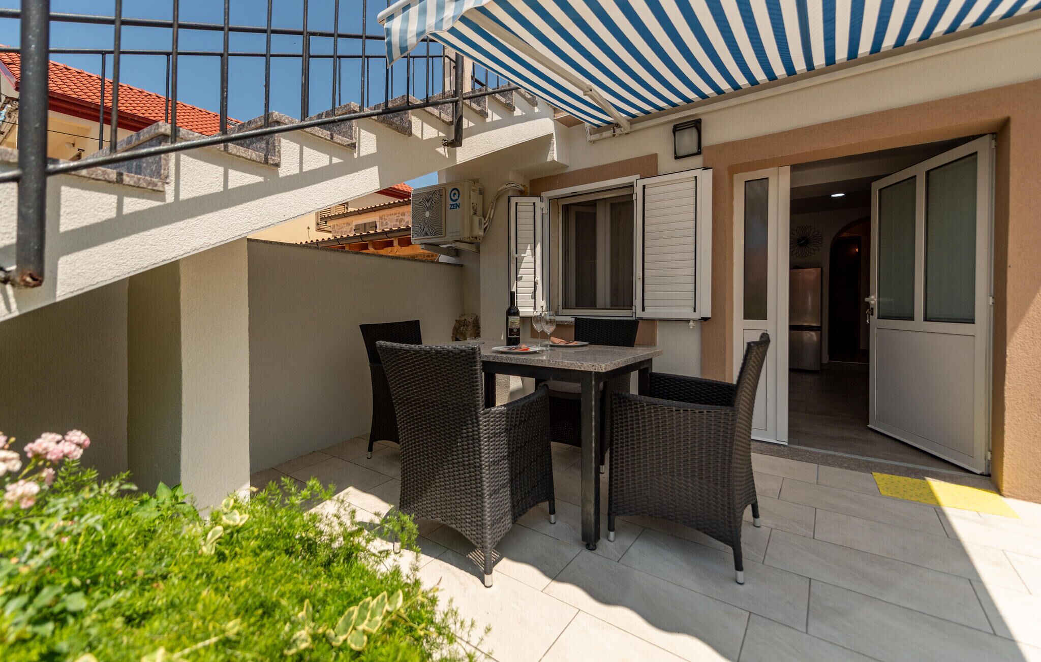 Een terras met een tafel en stoelen, een overdekt gedeelte met een gestreept zonnescherm, en uitzicht op een gebouw met een rode pannendak.