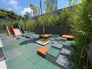 A rooftop garden with seating areas, a small tree, and a playground structure. - Innvista -Trends Brooklin. (São Paulo)