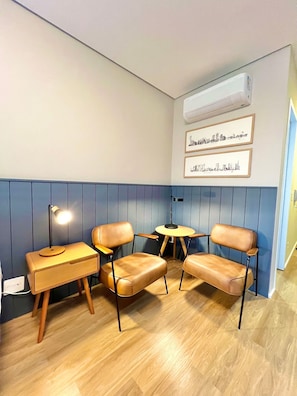 A room with a blue and white color scheme, featuring a wooden floor, a small round table, two leather chairs, a desk with a lamp, and framed cityscape pictures on the wall. - Innvista -Trends Brooklin. (São Paulo)