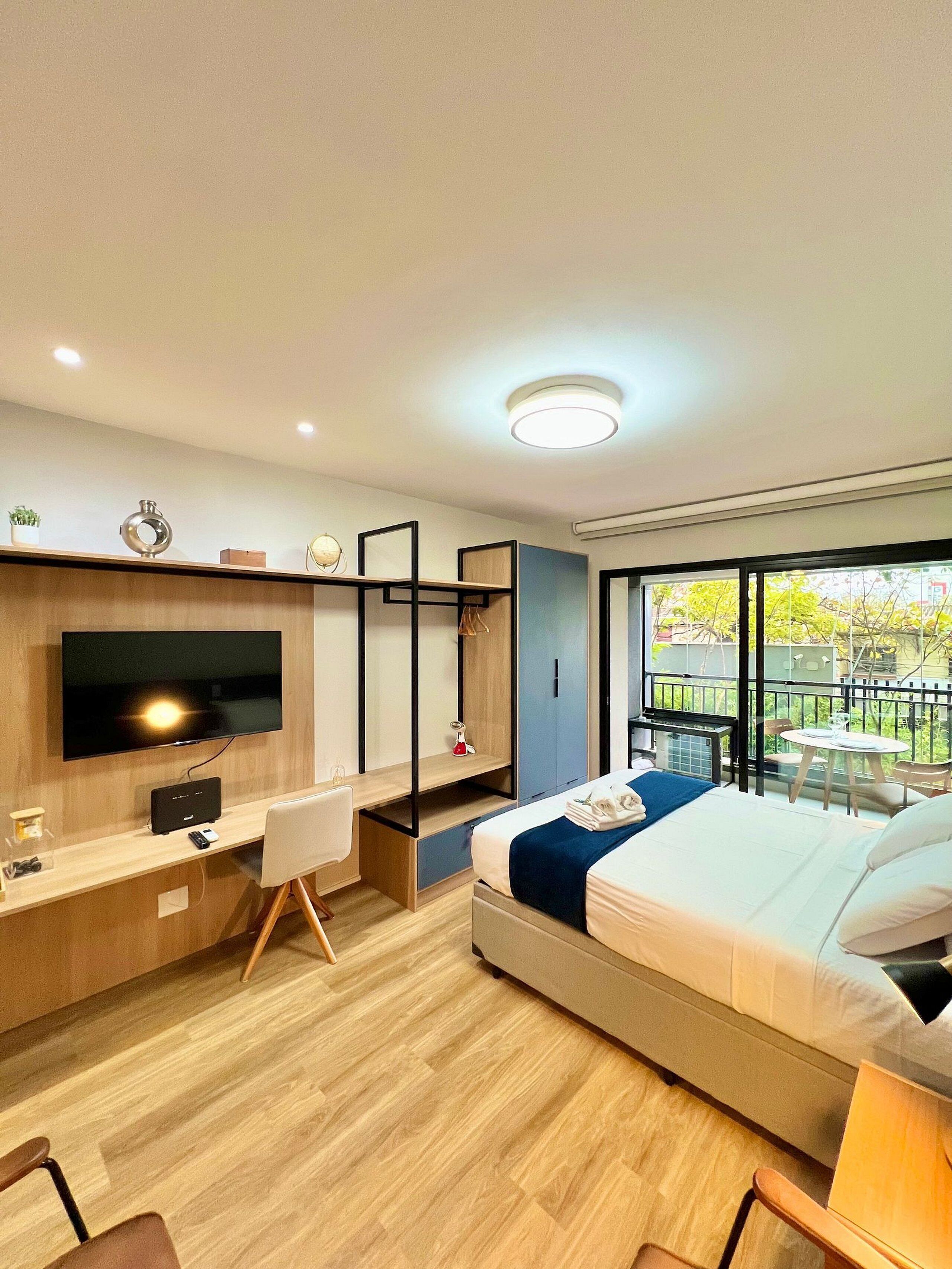 A modern hotel room with a large bed, a desk with a flat-screen TV, a small table, and a balcony with a view of greenery.