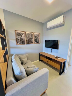 A modern living room with a grey sofa, a wooden TV stand, and three black and white cityscape paintings on the wall. - Innvista -Trends Brooklin. (São Paulo)