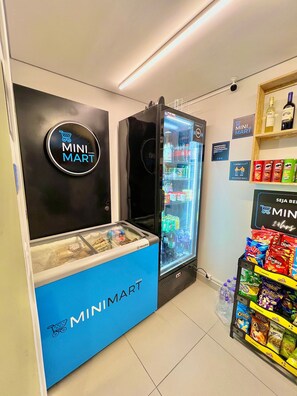 A Mini Mart kiosk with a refrigerated display and a freezer, surrounded by shelves stocked with snacks and beverages. - Innvista -Trends Brooklin. (São Paulo)