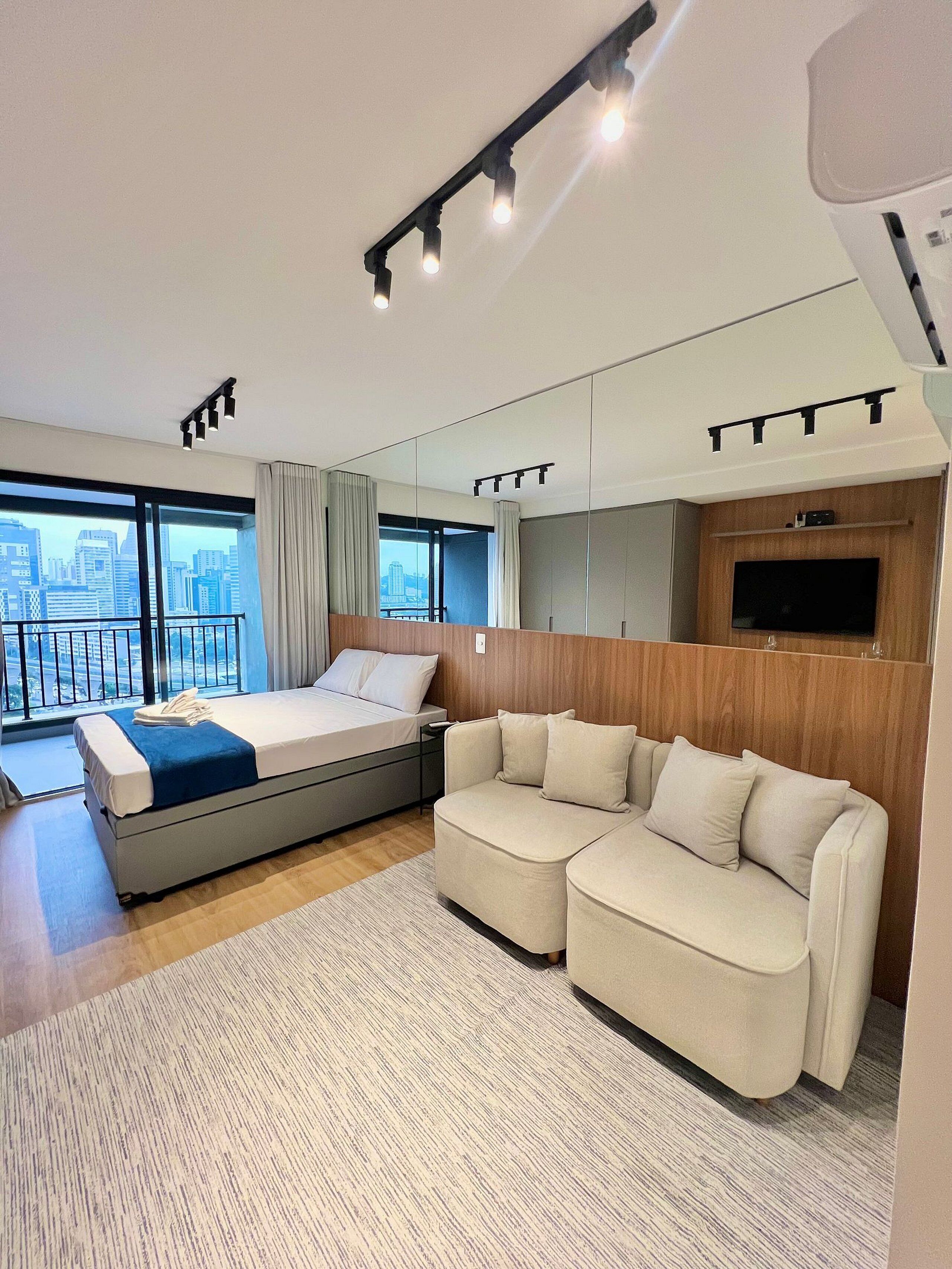 A modern hotel room with a large bed, a sofa, a TV, and a city view.