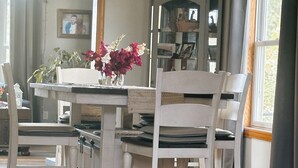 Un comedor con una mesa blanca, sillas y un florero. Hay una estantería y un espejo en la pared.
