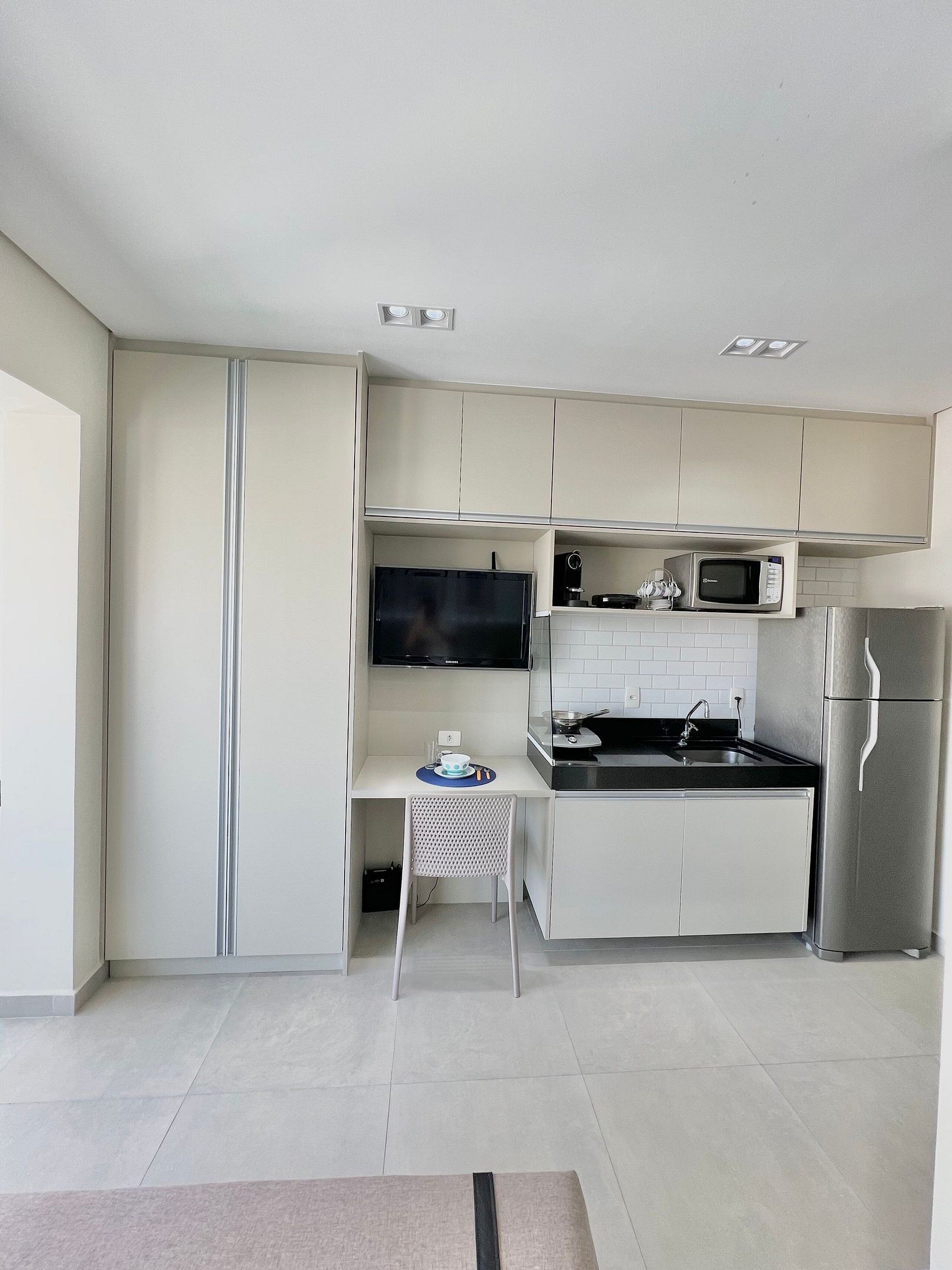 A modern kitchen with white cabinets, a stainless steel refrigerator, a microwave, and a flat-screen TV.