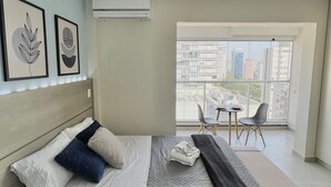 Et moderne hotelværelse med seng, et lille bord, to stole og et stort vindue med udsigt over byen.