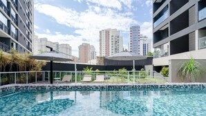 A swimming pool with tiled edges, surrounded by lounge chairs and umbrellas, with high-rise buildings in the background. - VN Quatá (São Paulo)