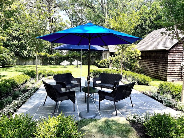 A patio with four wicker chairs and a table, a blue umbrella, and a wooden building in the background. - Wainscott Woods Retreat (Wainscott)