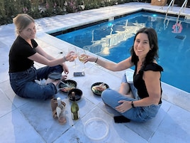 Two people sitting by a poolside with a dining setup including plates, a bottle, and a wine glass.