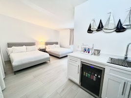 A hotel room with two beds, a kitchenette, a refrigerator, and a wall-mounted shelf with bottles.