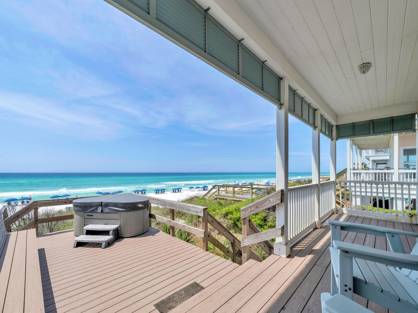 A wooden deck with a hot tub, overlooking a beach with umbrellas and a clear blue sky.