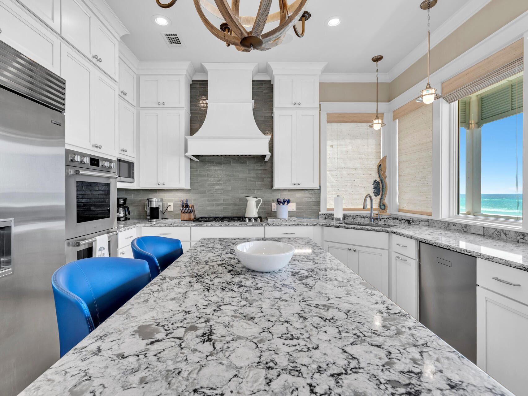 A modern kitchen with white cabinetry, a marble island, and a view of the ocean.