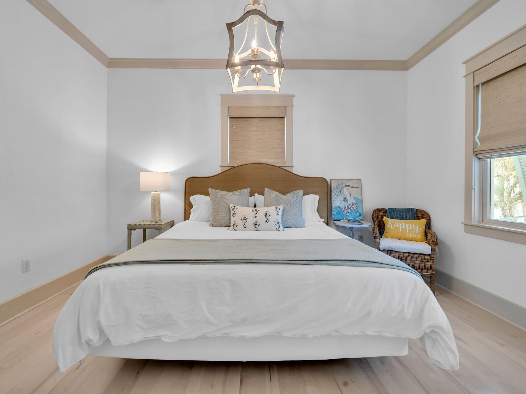 Een slaapkamer met een groot bed, een nachtkastje met lamp, een rieten stoel, een raam met zonwering en een plafondlamp.
