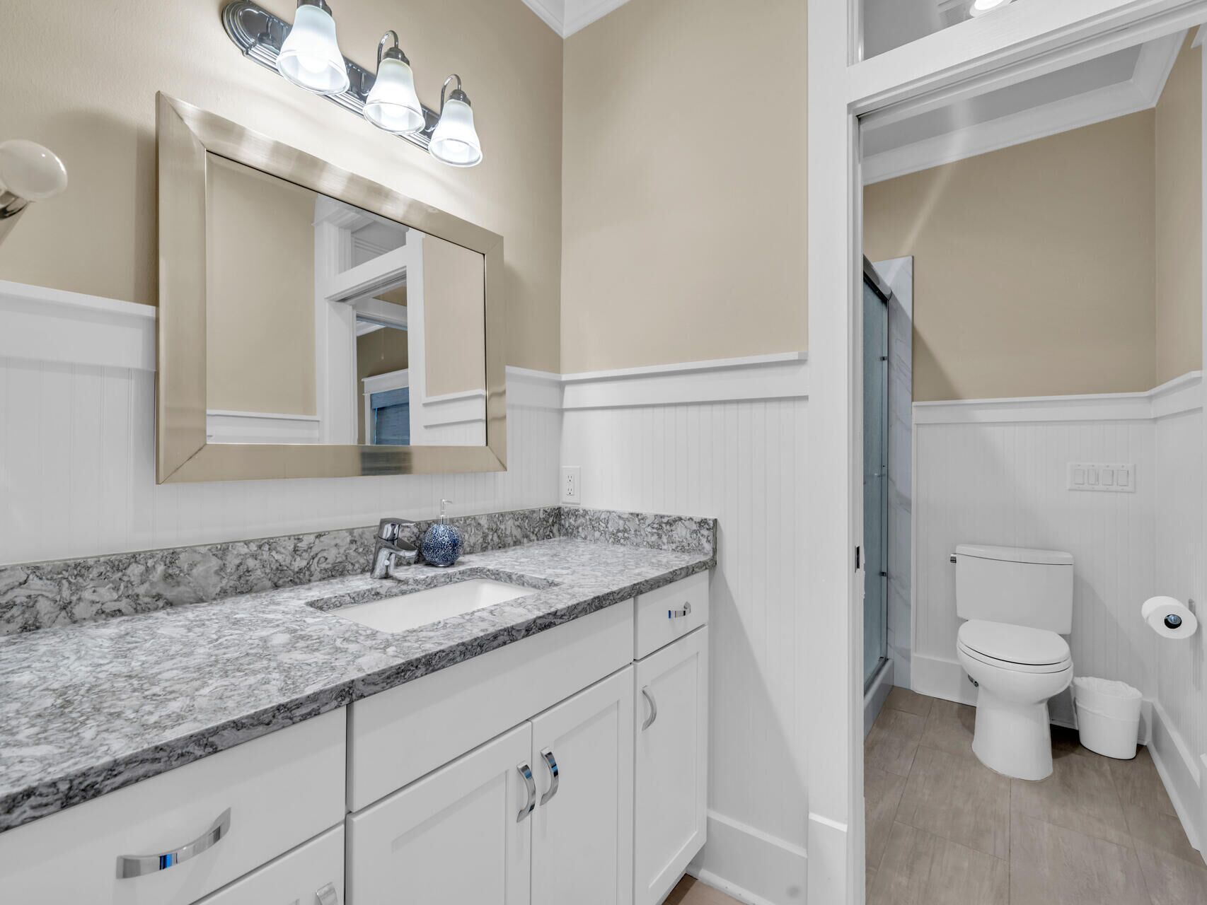 A bathroom with a marble countertop, a sink, a toilet, and a mirror with lights above it.