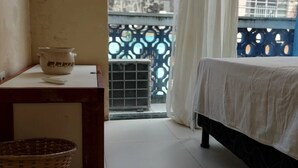 Una habitación con cama, una mesa con jarra de cerámica, una cesta y ventana con vistas a una valla azul y unidad de aire acondicionado.