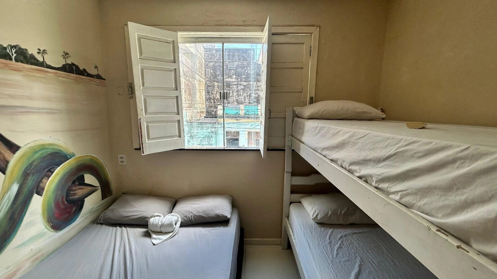 Un alojamiento pequeño y con una sola habitación, con literas, una ventana con contraventanas abiertas y un mural en la pared.