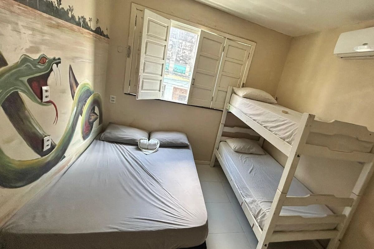 Um quarto com beliche, um mural na parede, uma janela com as persianas abertas e um aparelho de ar condicionado.