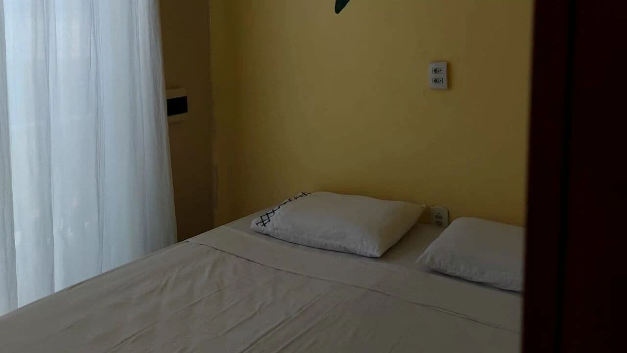 Una habitación sencilla con cama, ropa de cama blanca, una pared amarilla lisa y una ventana con cortinas blancas.