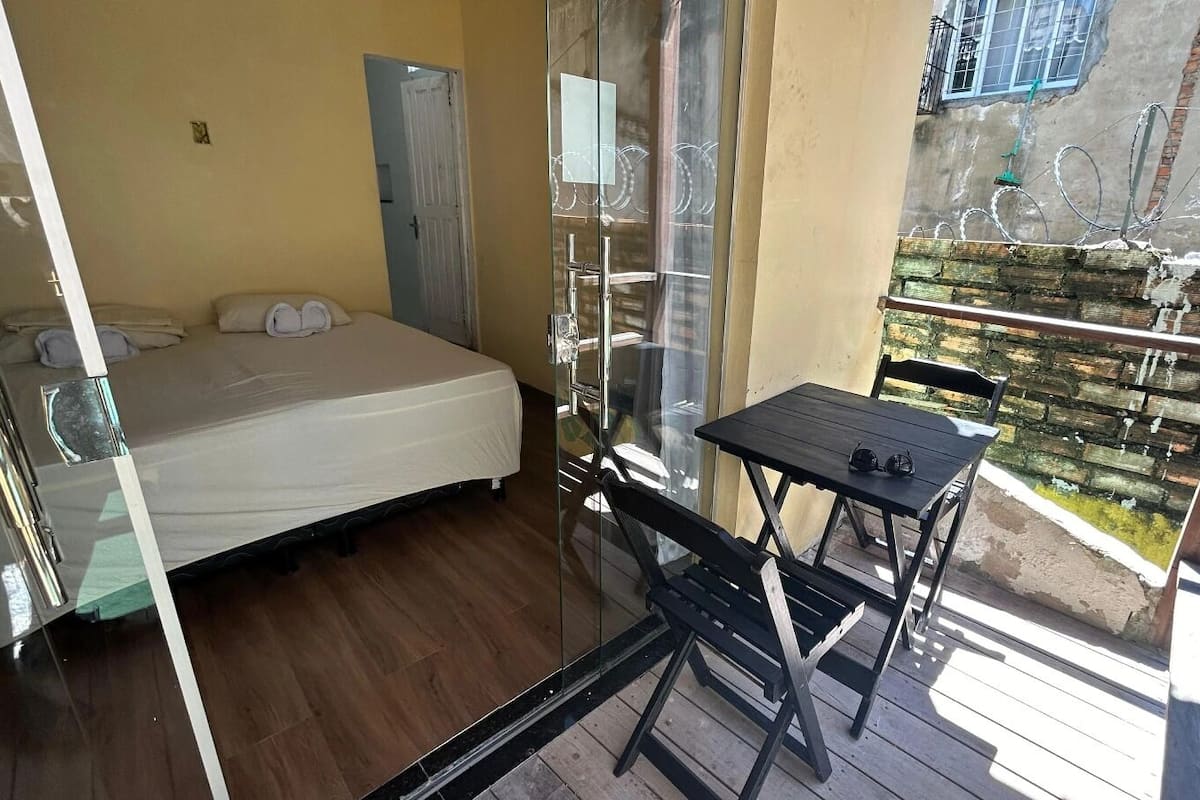 Quarto pequeno com cama, mesa e cadeira. Há uma janela e uma porta.
