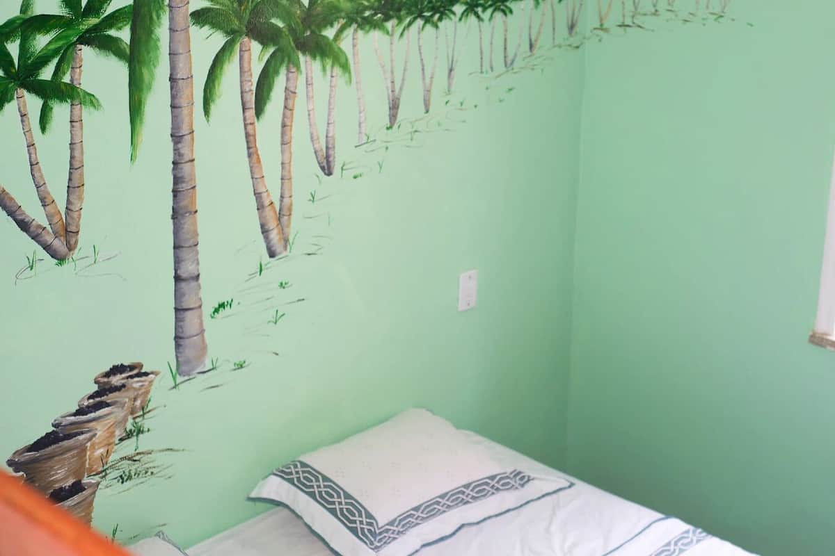 Quarto com parede verde, cama com travesseiro estampado e um mural de palmeiras.
