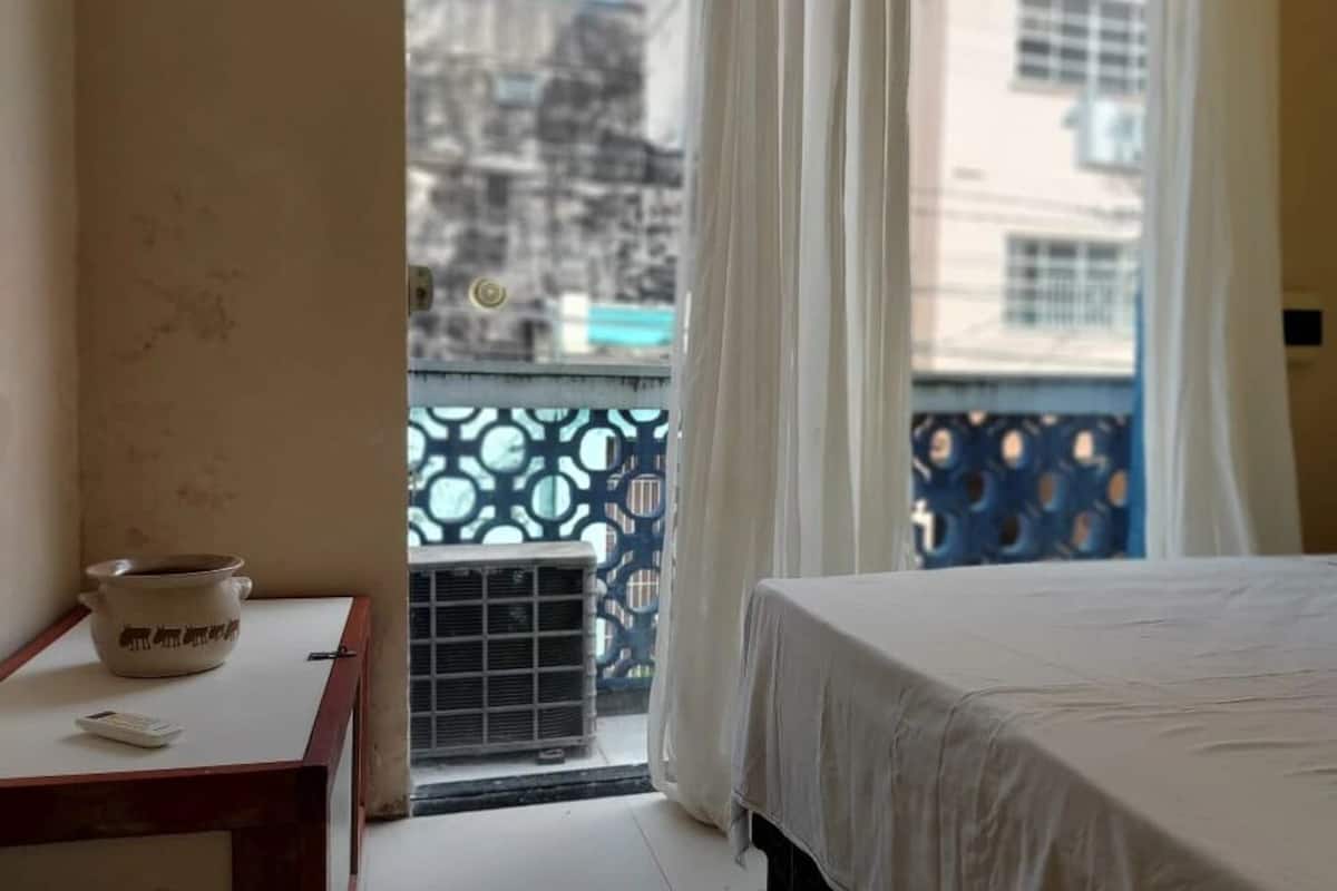 Quarto de hotel com cama, uma pequena mesa com chaleira e controle remoto, e janela com vista para edifícios e varanda com grade estampada.