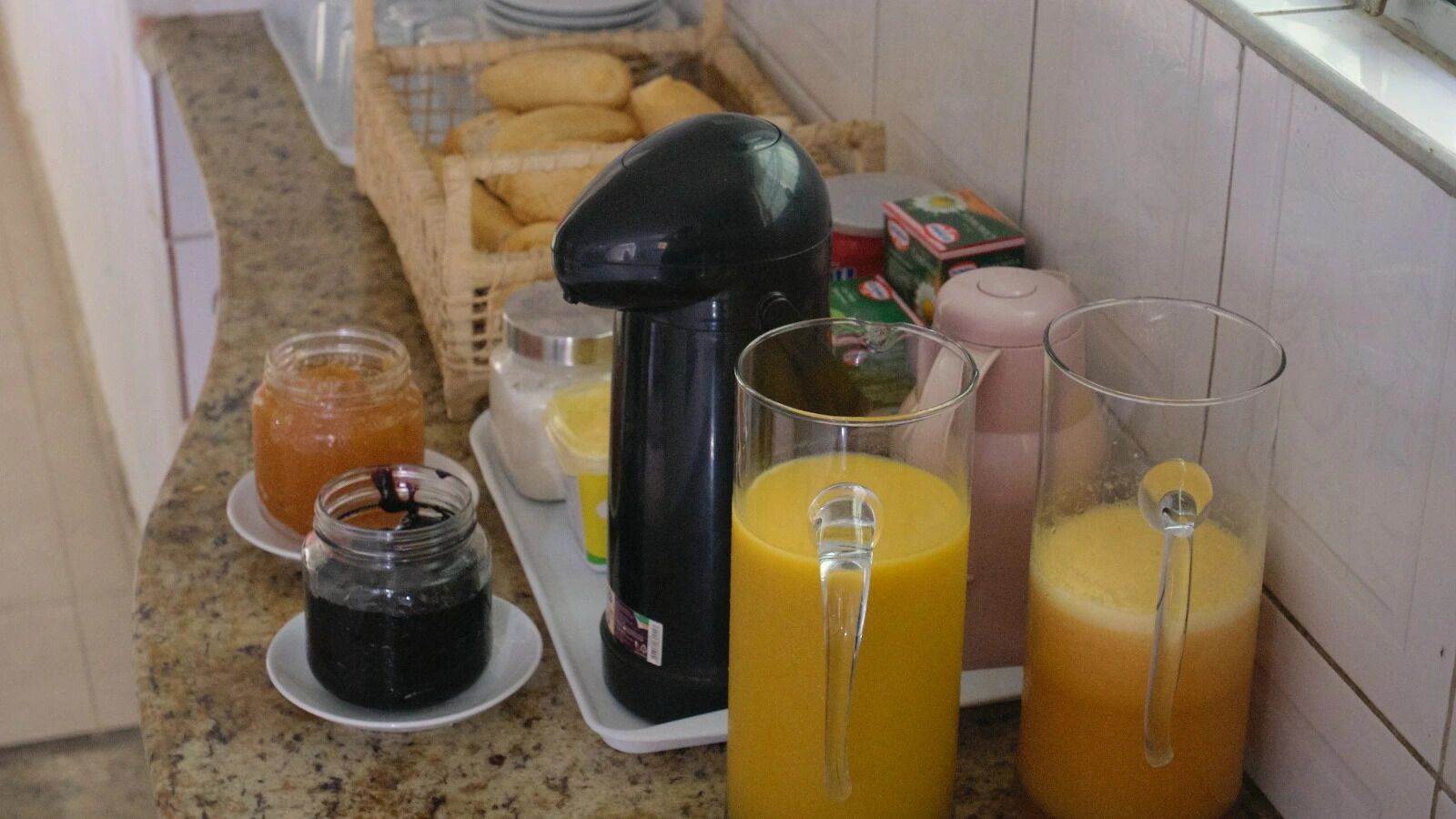 Una encimera de cocina con diversos alimentos y bebidas, incluyendo frascos de conservas, una cafetera y vasos de zumo de naranja.