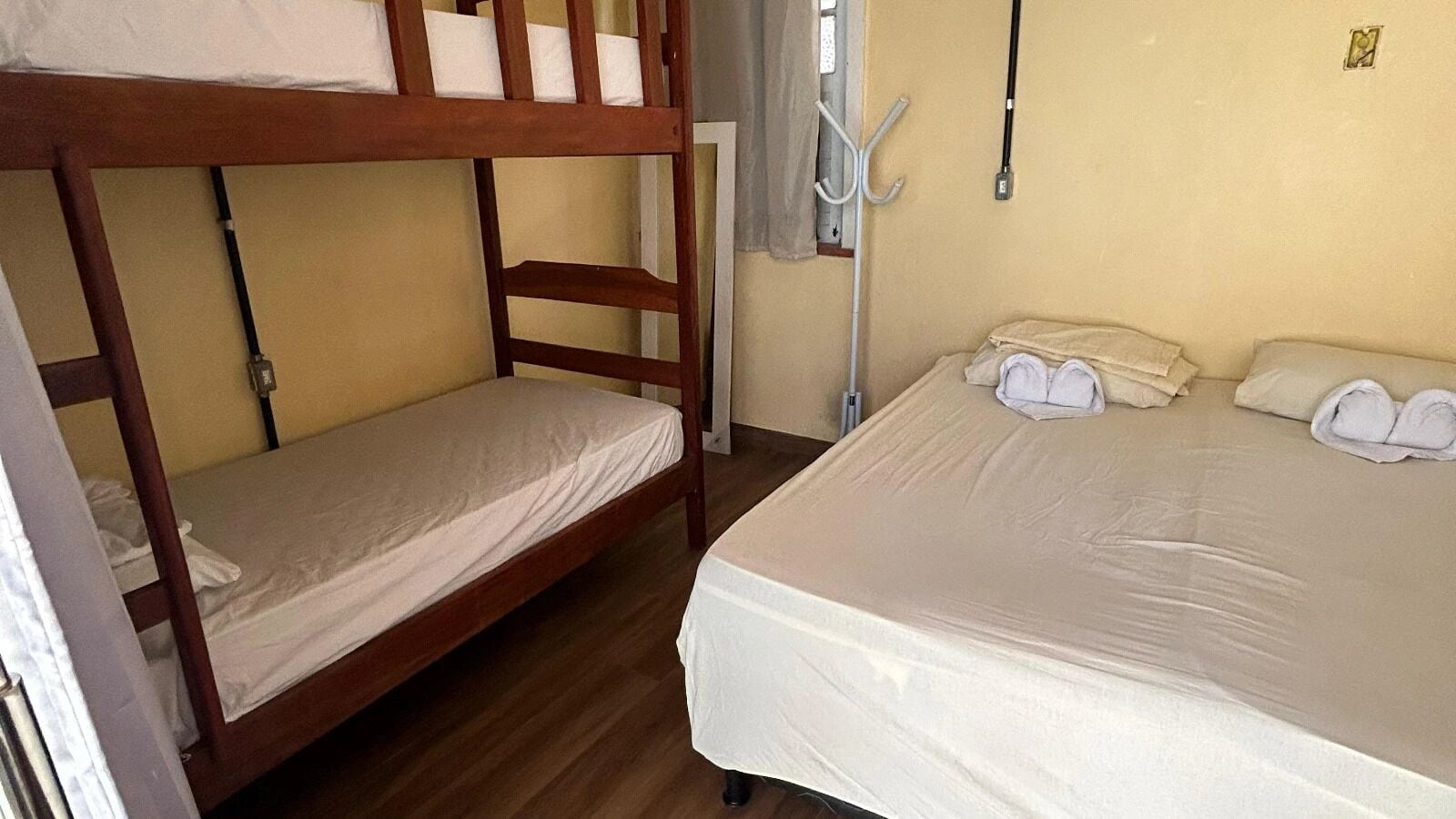Habitación con dos camas, una litera y una individual, ambas con ropa blanca. Hay una ventana con cortina y un perchero con un abrigo colgado.