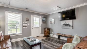 A living room with a brown leather couch, a glass coffee table, a wooden bench, a television mounted on the wall, and a large window with blinds.