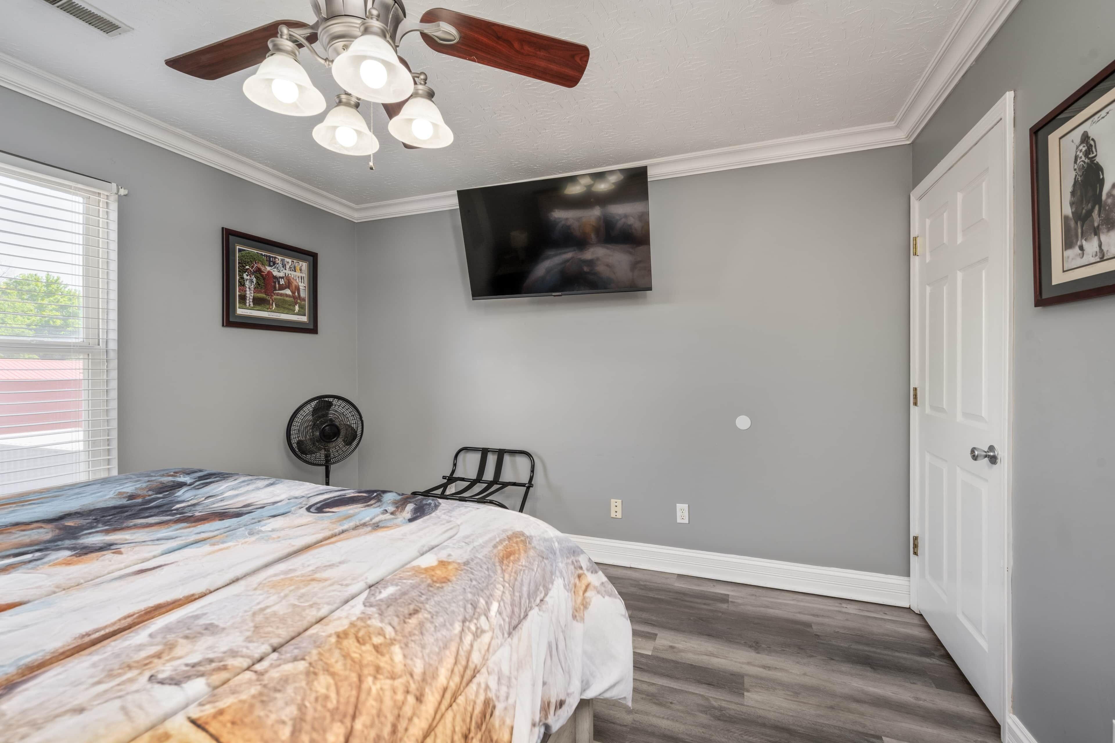 Une chambre à coucher avec un lit, un ventilateur de plafond, un téléviseur fixé au mur et une porte.