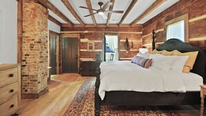 Un dormitorio con vigas de madera a la vista, pared de ladrillo, una cama grande con muchas almohadas, una mesita de noche, un aparador y un ventilador de techo.