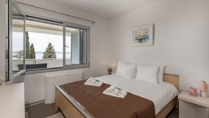 Quarto de hotel com cama, janela com vista para árvores e um corpo d'água, um quadro na parede e uma mesa de cabeceira com abajur e um vaso.