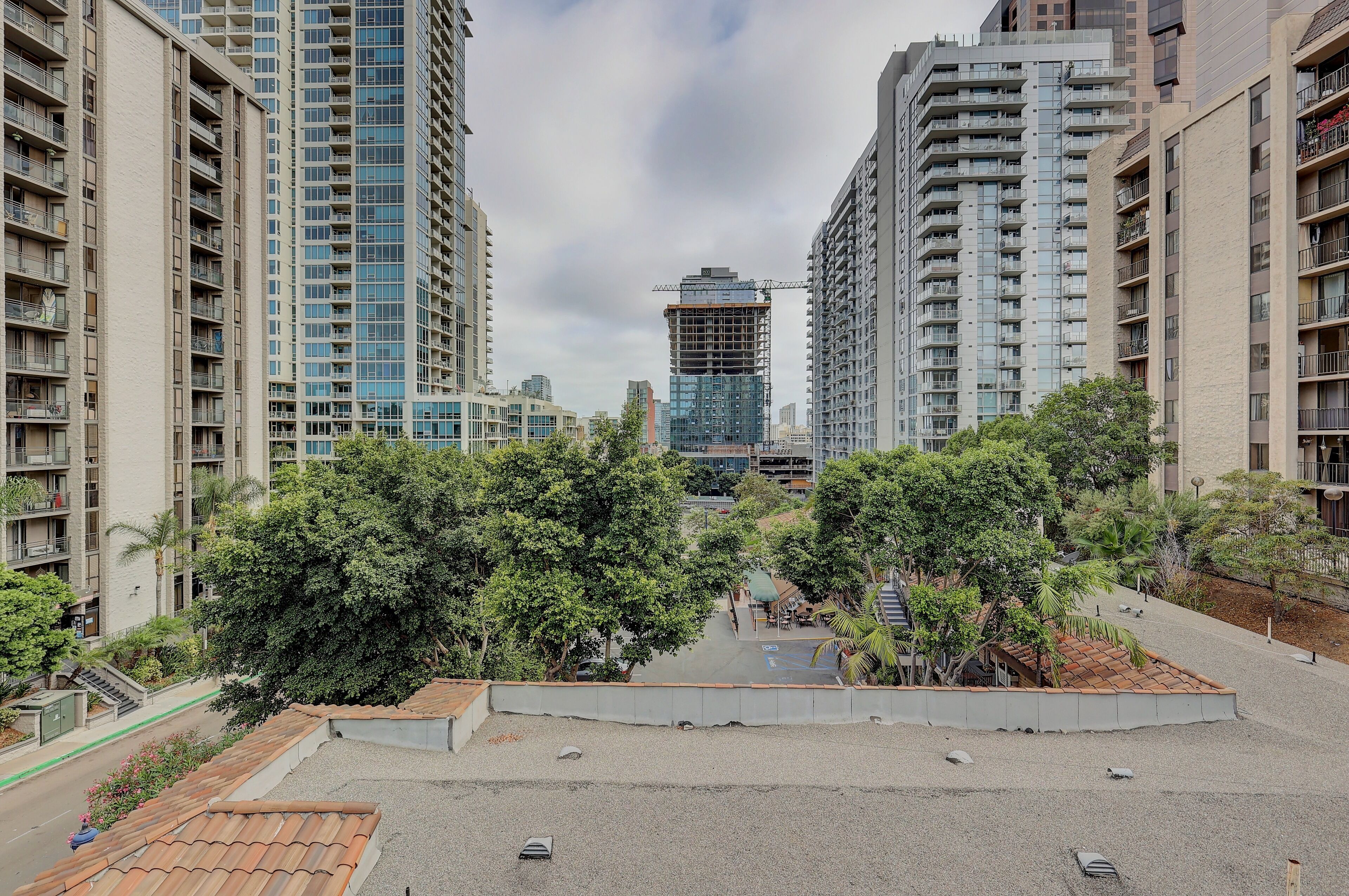Un edificio residencial alto con varios balcones, una obra en construcción al fondo y una zona ajardinada con árboles y un sendero pavimentado.
