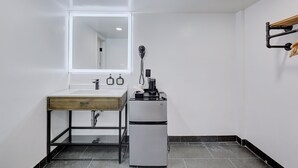 A compact bathroom with a white sink, a mirror, a small refrigerator, and a wooden shelf. - Ash&9th (San Diego)
