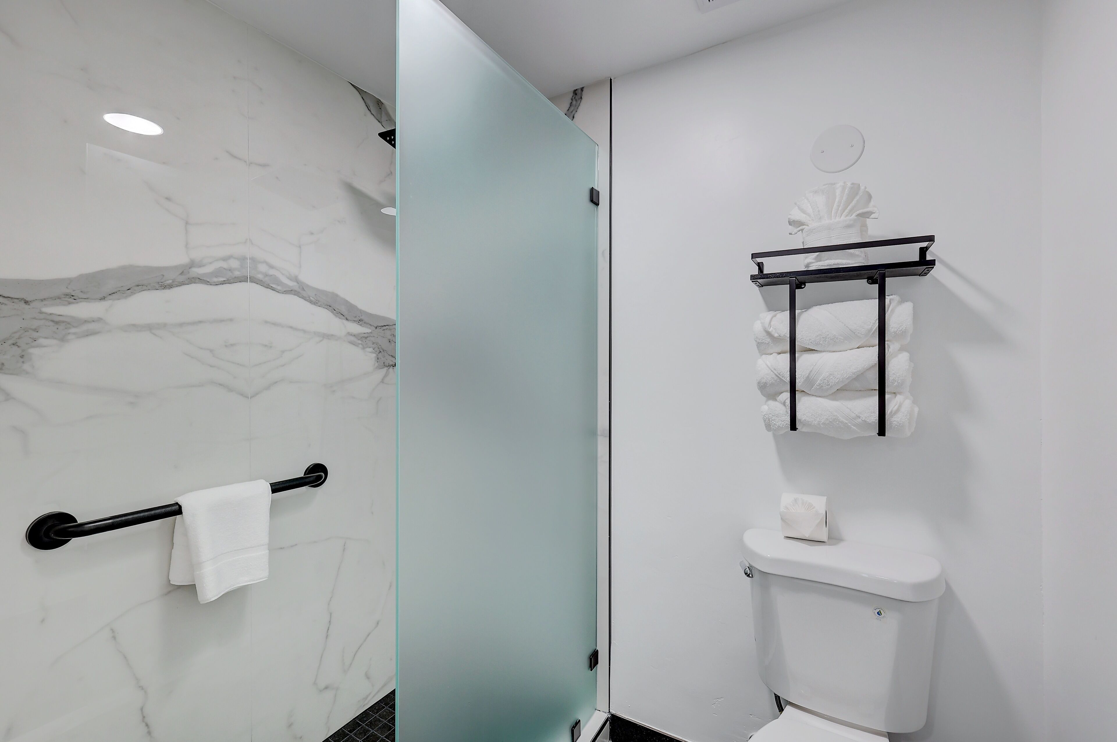 A bathroom with a marble wall, a glass shower door, a toilet with a towel rack above it, and a shelf with towels.