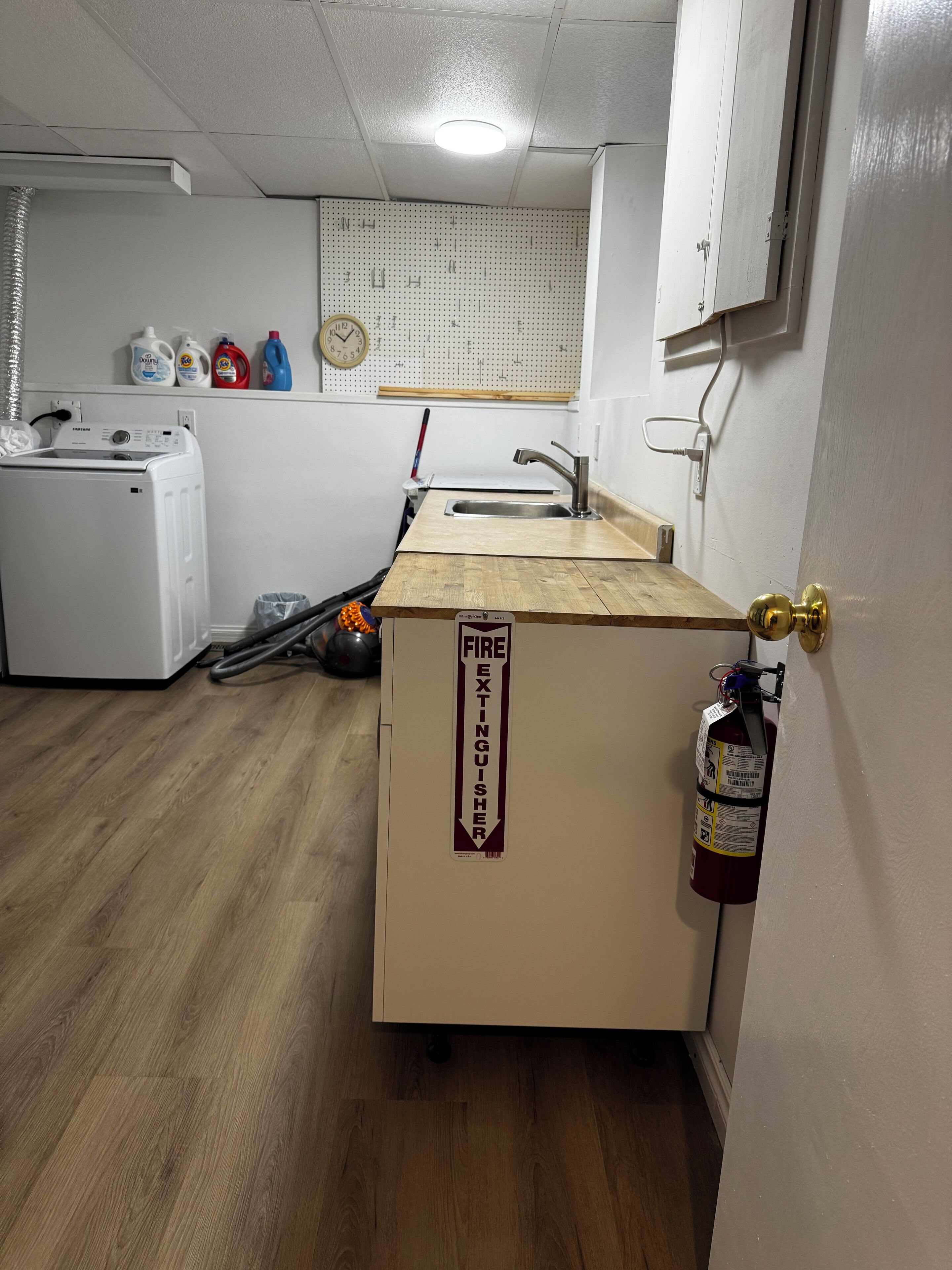 Una pequeña kitchenette con encimera de madera, un extintor y una lavadora al fondo.