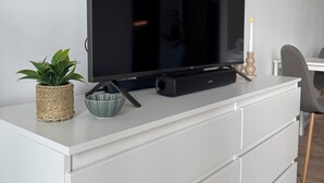 Litteänäyttöinen televisio on sijoitettu valkoiselle lipastolle, jonka päällä on punottu kori, lasikulho ja kynttilä.