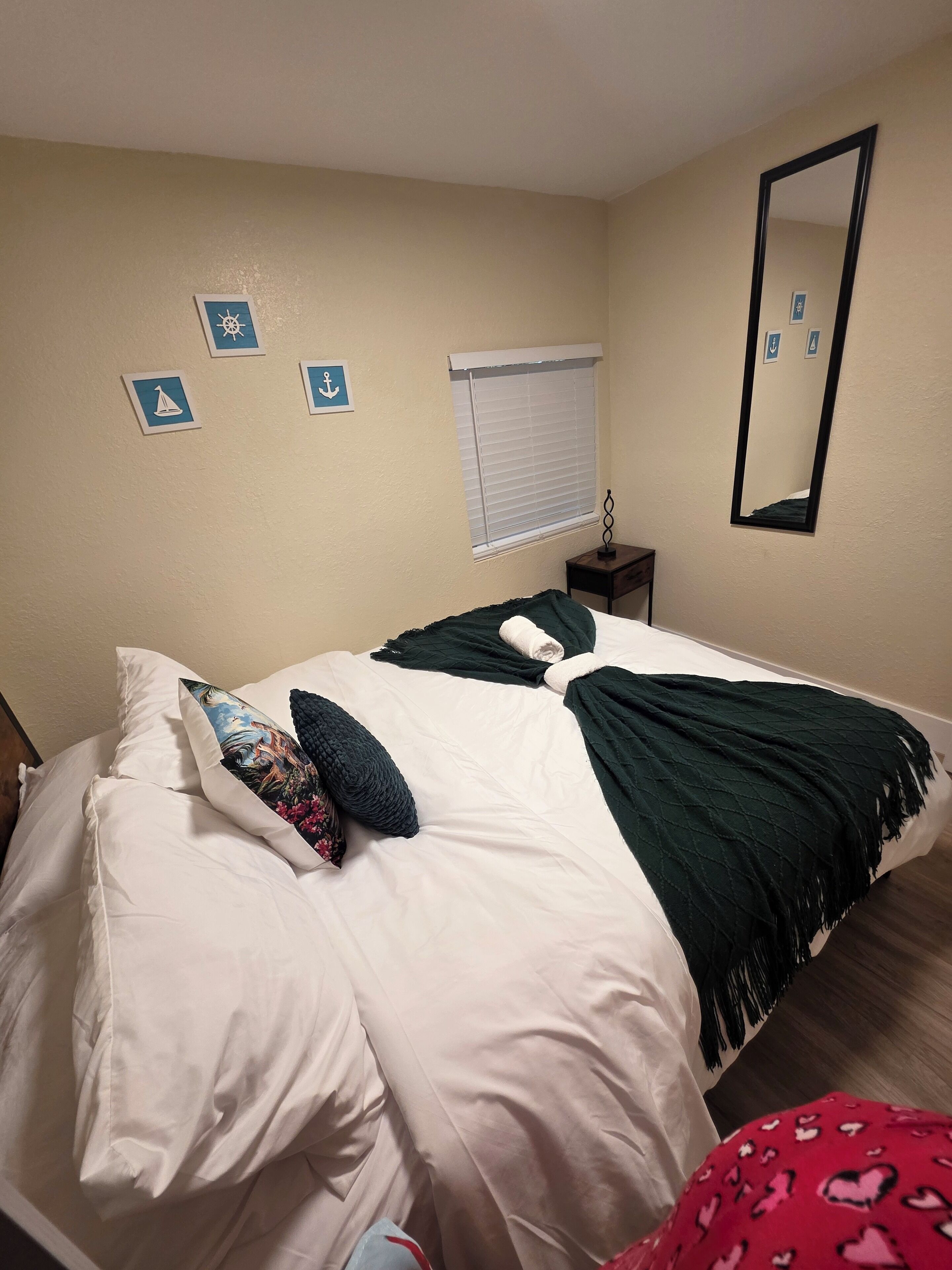 Un lit bien fait, recouvert d’une couverture verte et agrémenté d’un coussin décoratif, une petite table de chevet avec une lampe, et une fenêtre avec des stores.