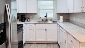 Una cocina moderna con armarios blancos, un refrigerador de acero inoxidable, horno empotrado, un lavamanos con grifo y una ventana con persianas.