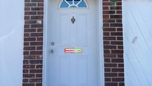 Una puerta blanca con el número "32" arriba, una pequeña ventana con diseño en forma de abanico y un buzón verde en la pared de ladrillo.