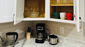 Una cocina con armarios blancos, cafetera, tetera y diversos vasos en estantes.