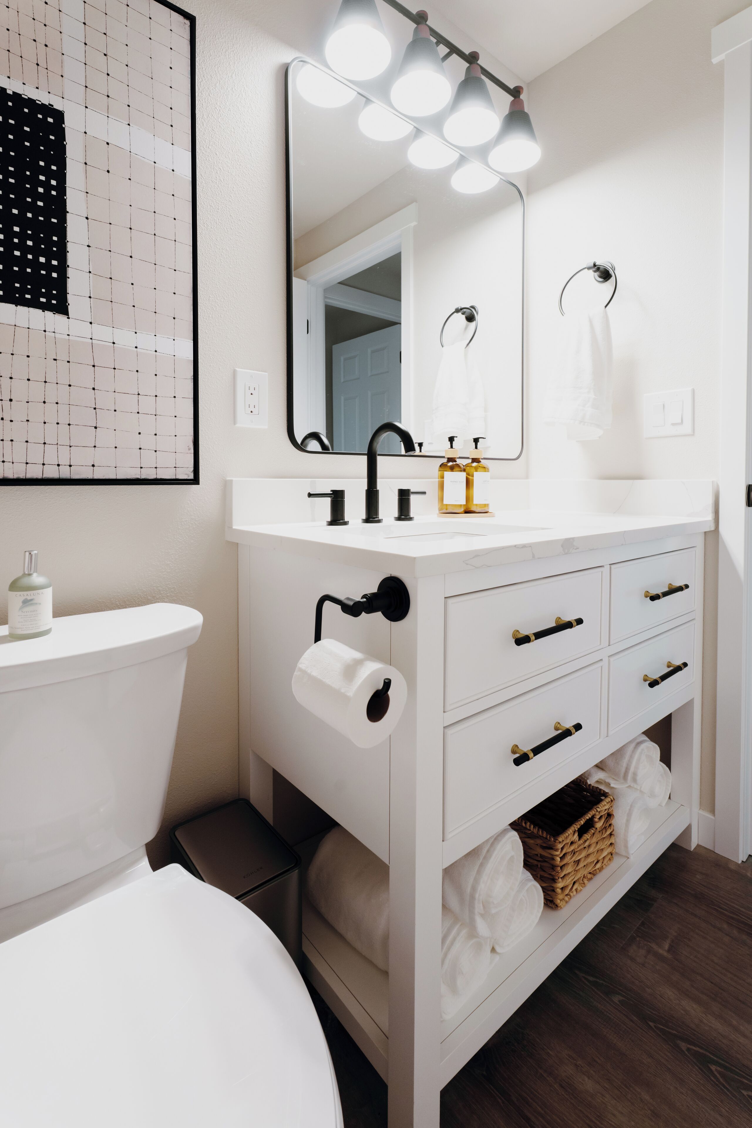Un baño moderno con tocador blanco, un espejo grande iluminado, inodoro y una obra de arte enmarcada en la pared.