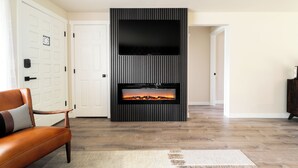 A modern living room with a fireplace, a flat-screen TV mounted above it, and a leather sofa. - Gas Fire Pit/Coffee Bar/Walk to Downtown/EV/Pets/AC (Enumclaw)