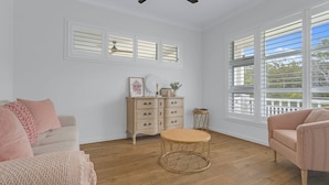 A spacious room with a large window, a wooden floor, a beige sofa, a round wicker table, and a white dresser with drawers. - Bikinis & Martinis - Jervis Bay (Vincentia)