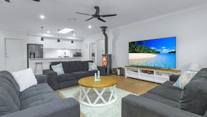A modern living room with grey sofas, a wooden coffee table, and a large flat-screen TV displaying a beach scene. - Bikinis & Martinis - Jervis Bay (Vincentia)