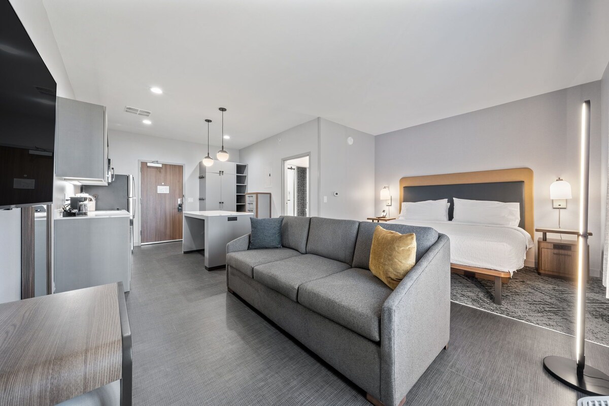 Een moderne hotelkamer met een grijze bank, een bed met witte lakens, een klein bureau en een kitchenette.