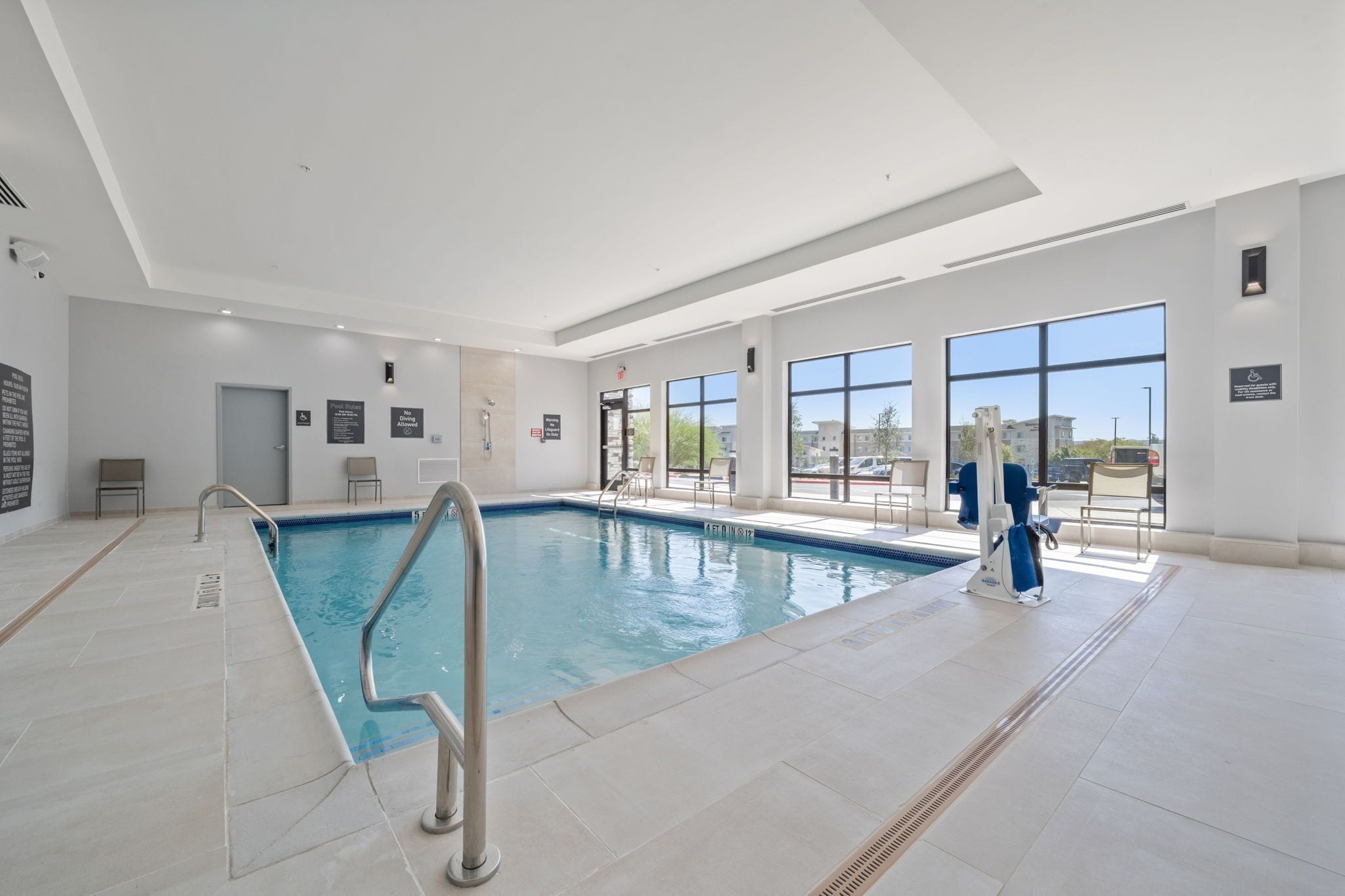 An indoor swimming pool with clear glass walls, a tiled floor, and a few chairs and a changing area.